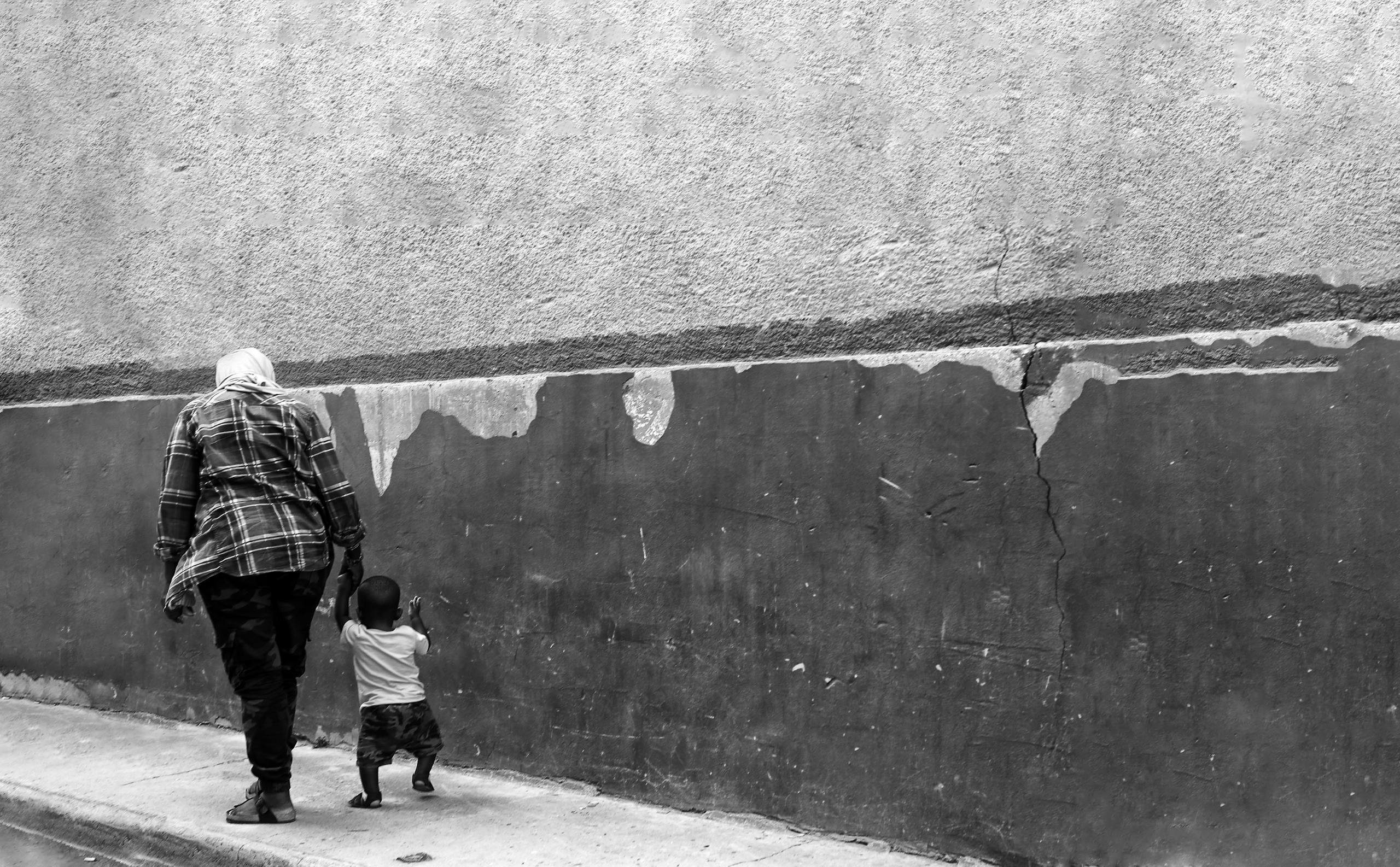 A mother and child walking beside a cracked urban wall in black and white.